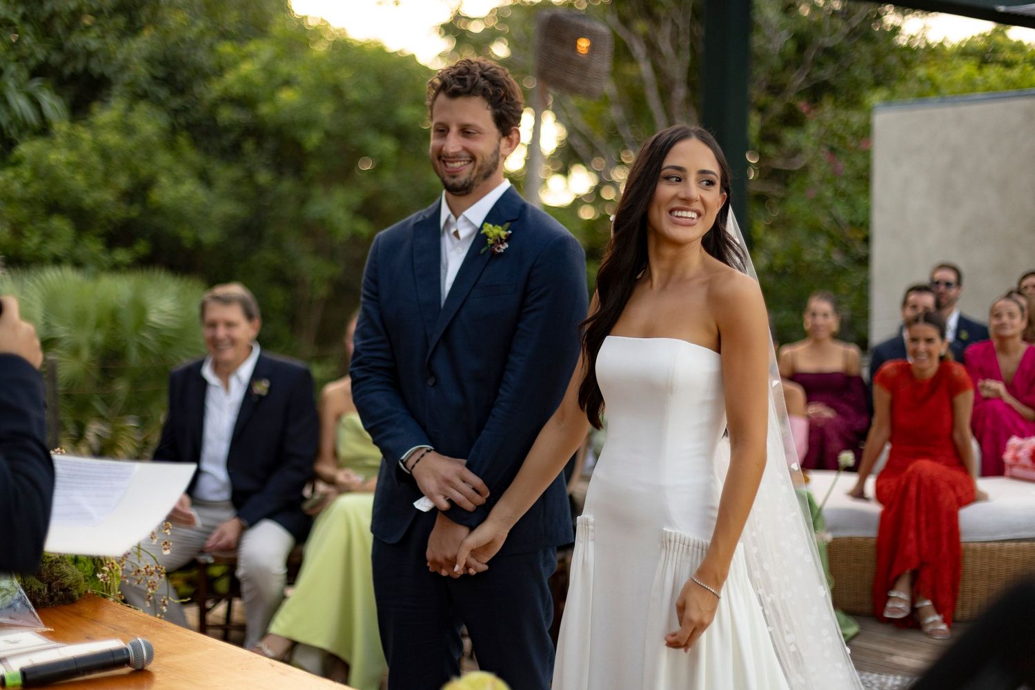 Smiling bride and groom hold hands during outdoor wedding ceremony surrounded by lush greenery.