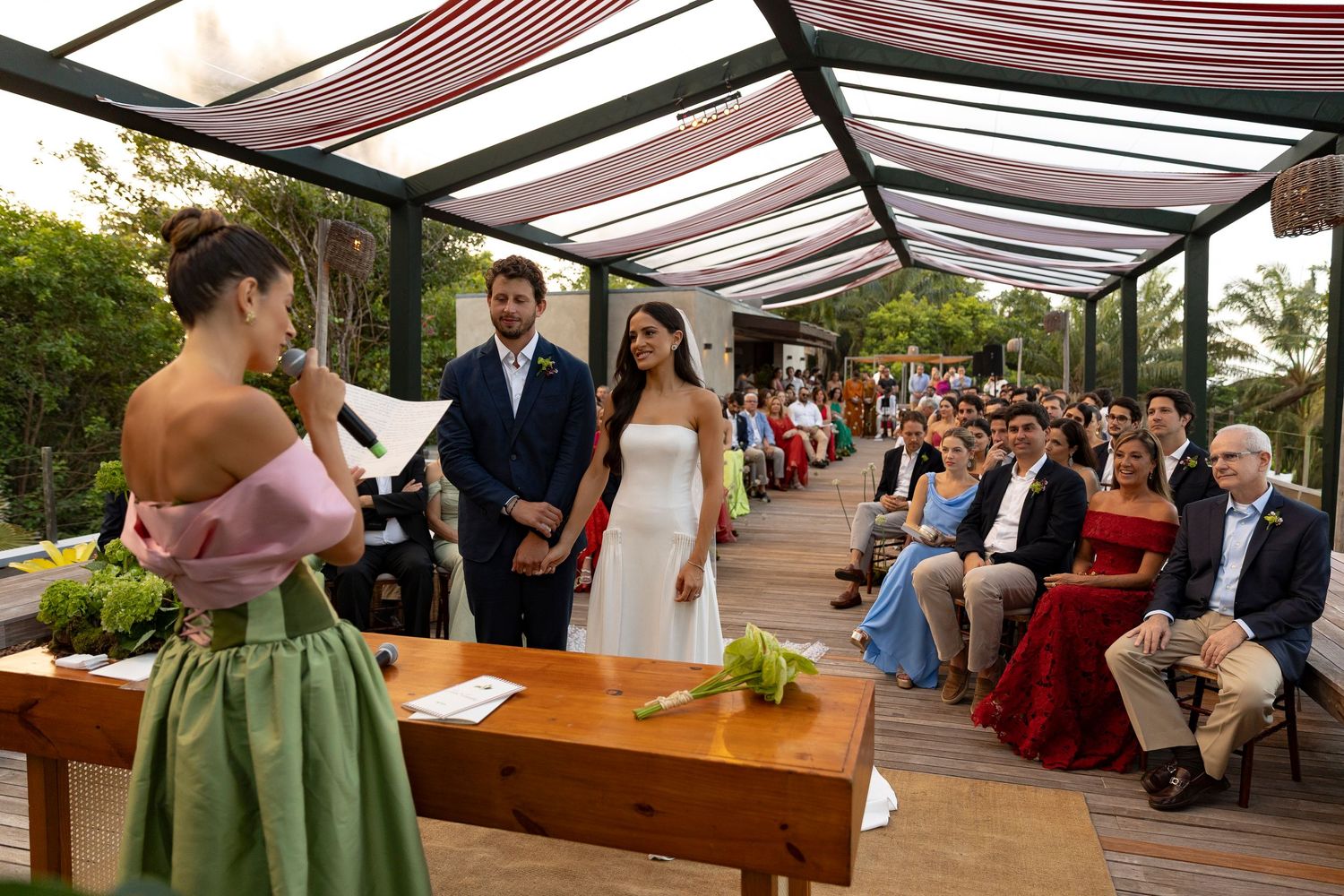Officiant reads vows at outdoor wedding ceremony under pergola with seated guests in background.