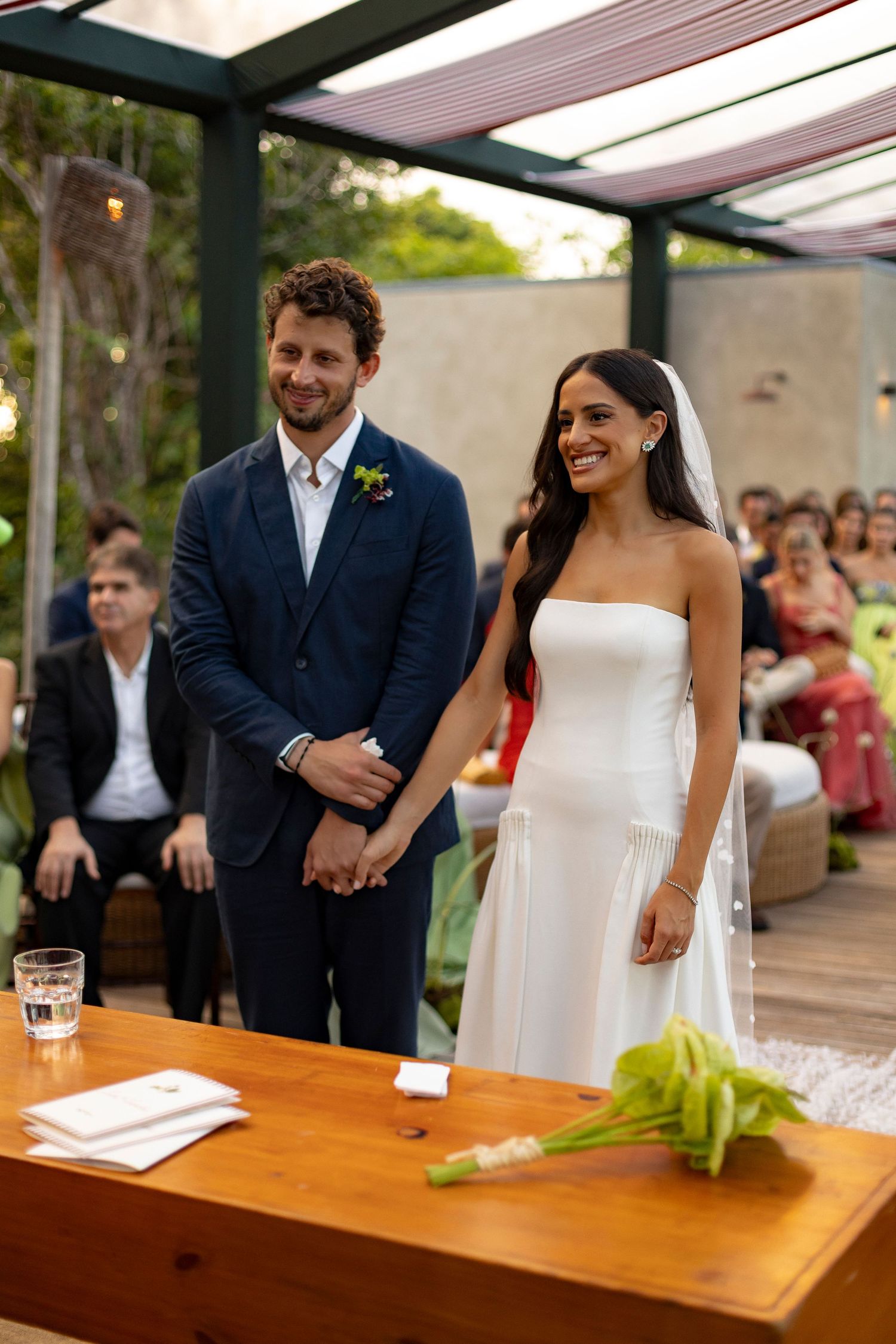 Couple smiling at outdoor wedding ceremony, groom in navy suit, bride in strapless white gown with veil.