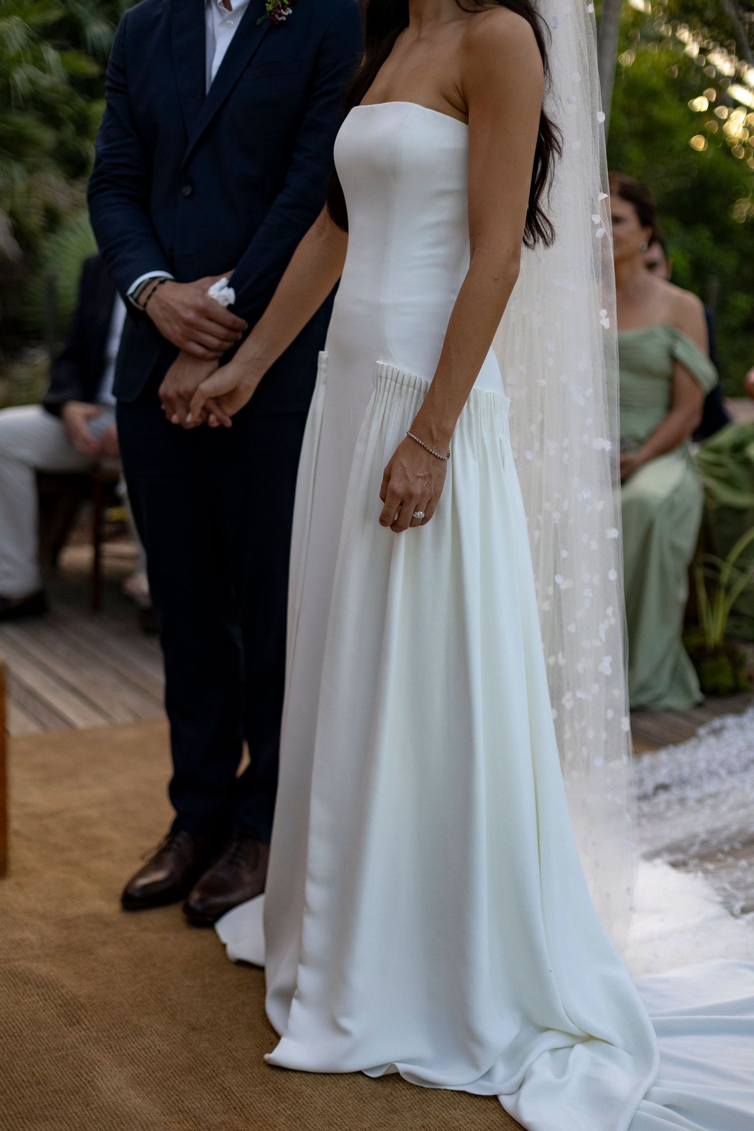 Bride in strapless white gown holds hands with groom in navy suit during outdoor wedding ceremony.