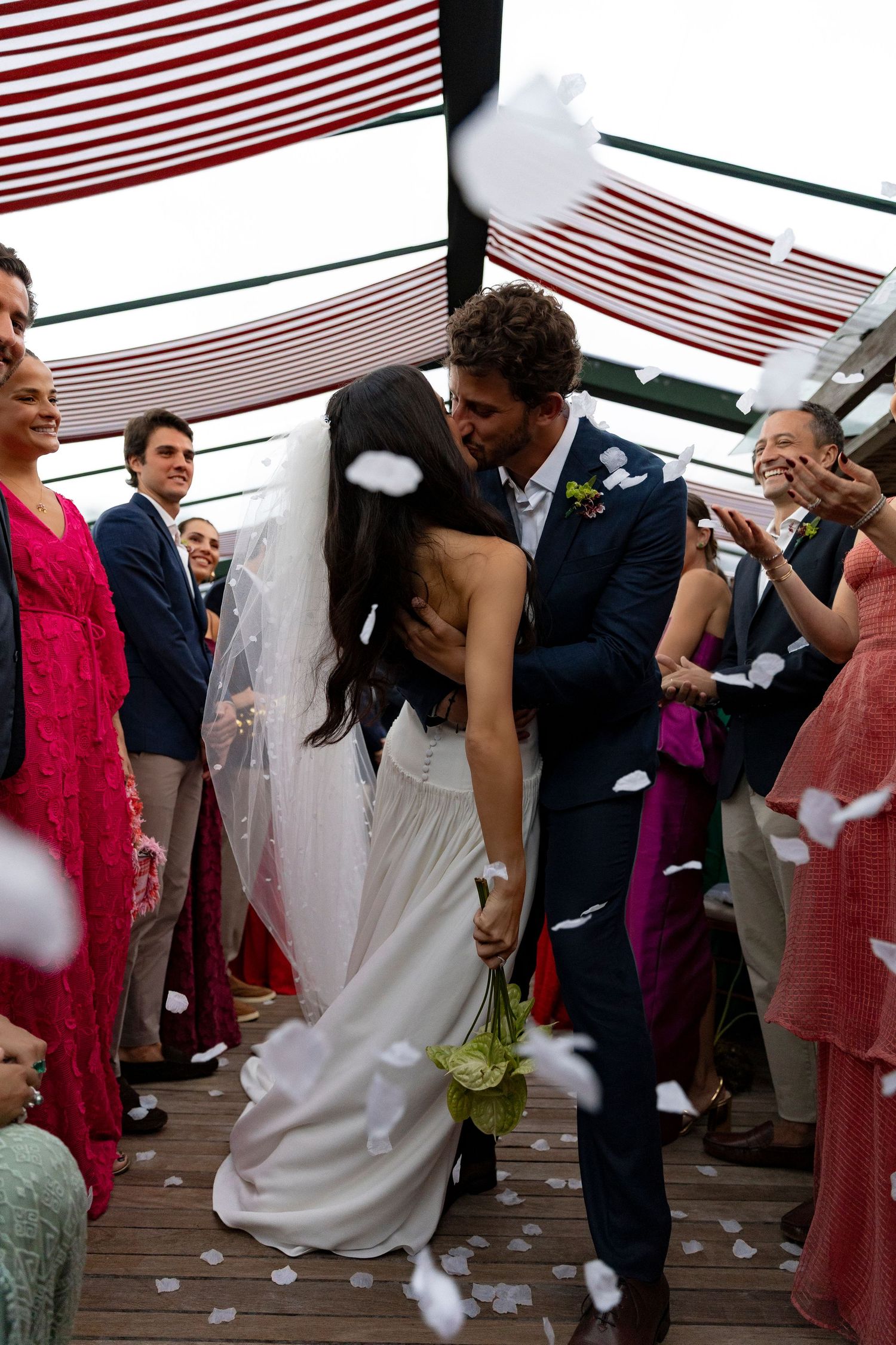 Bride and groom share a first kiss at outdoor wedding ceremony as guests toss white flower petals in celebration.