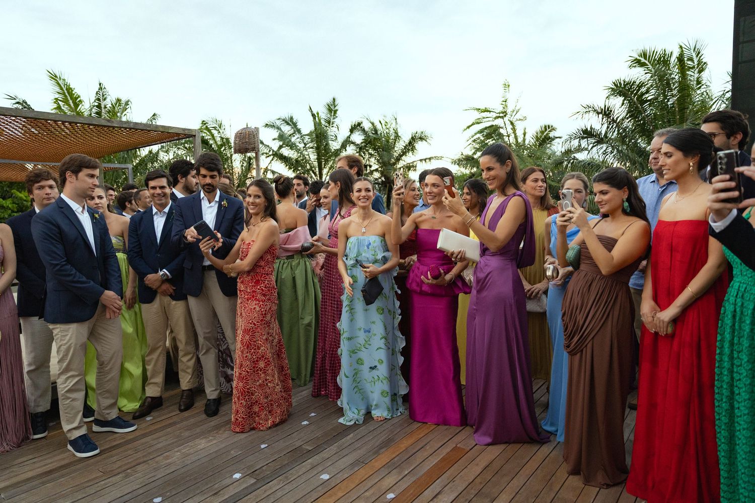 Elegantly dressed wedding guests in colorful gowns and suits gathered on a tropical outdoor venue deck with palm trees.