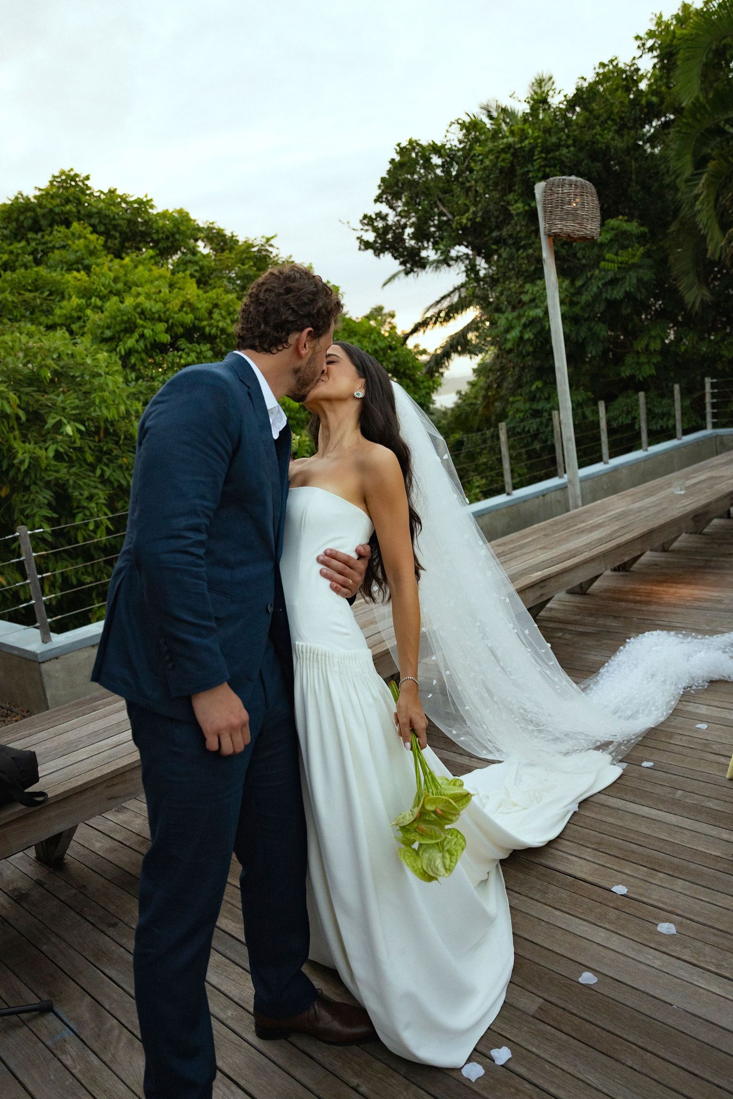 Newlywed couple sharing a kiss on a wooden deck, bride in white gown with veil, groom in navy suit.