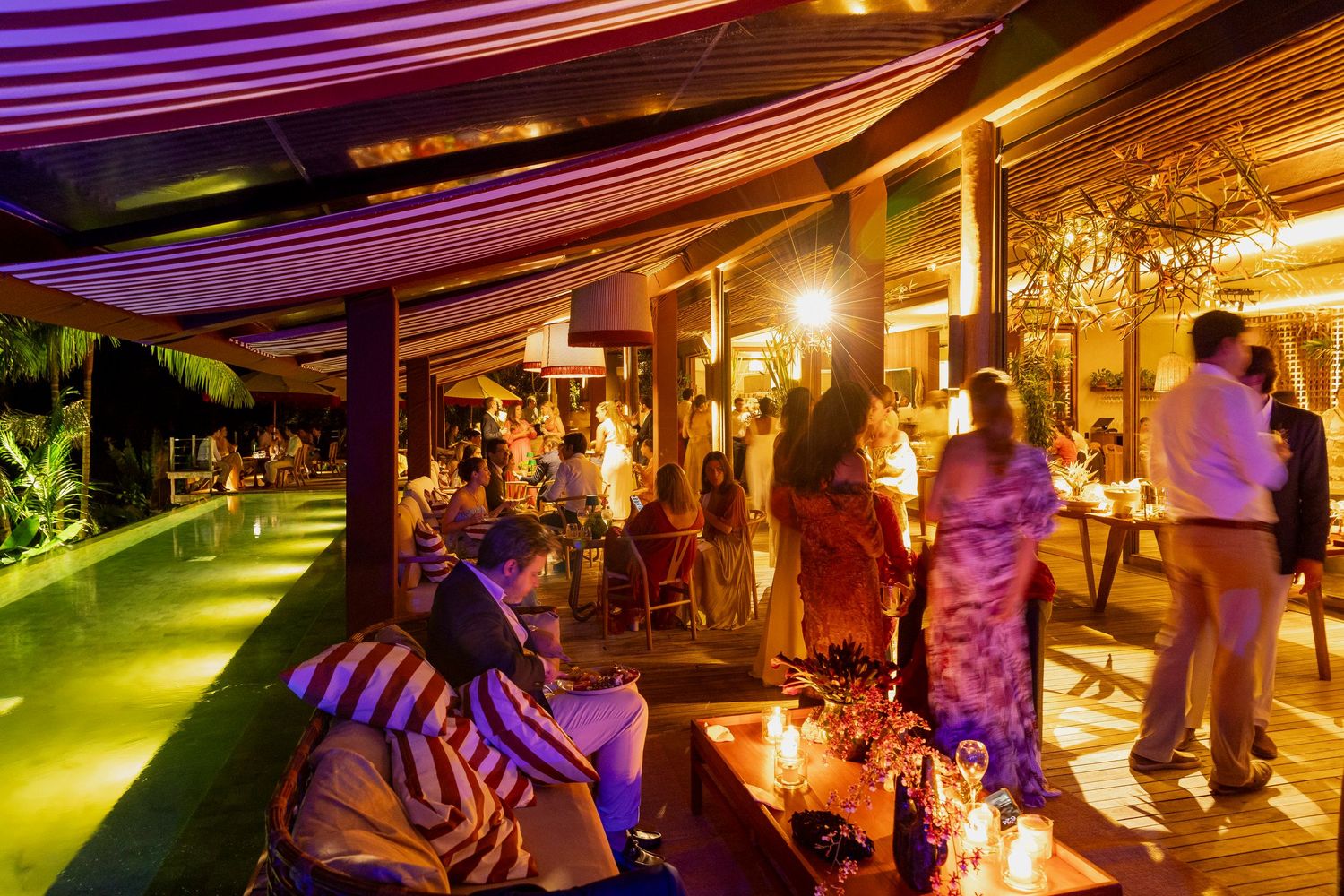 Elegant outdoor evening party with guests socializing near a pool, illuminated by warm golden lights under a striped canopy.