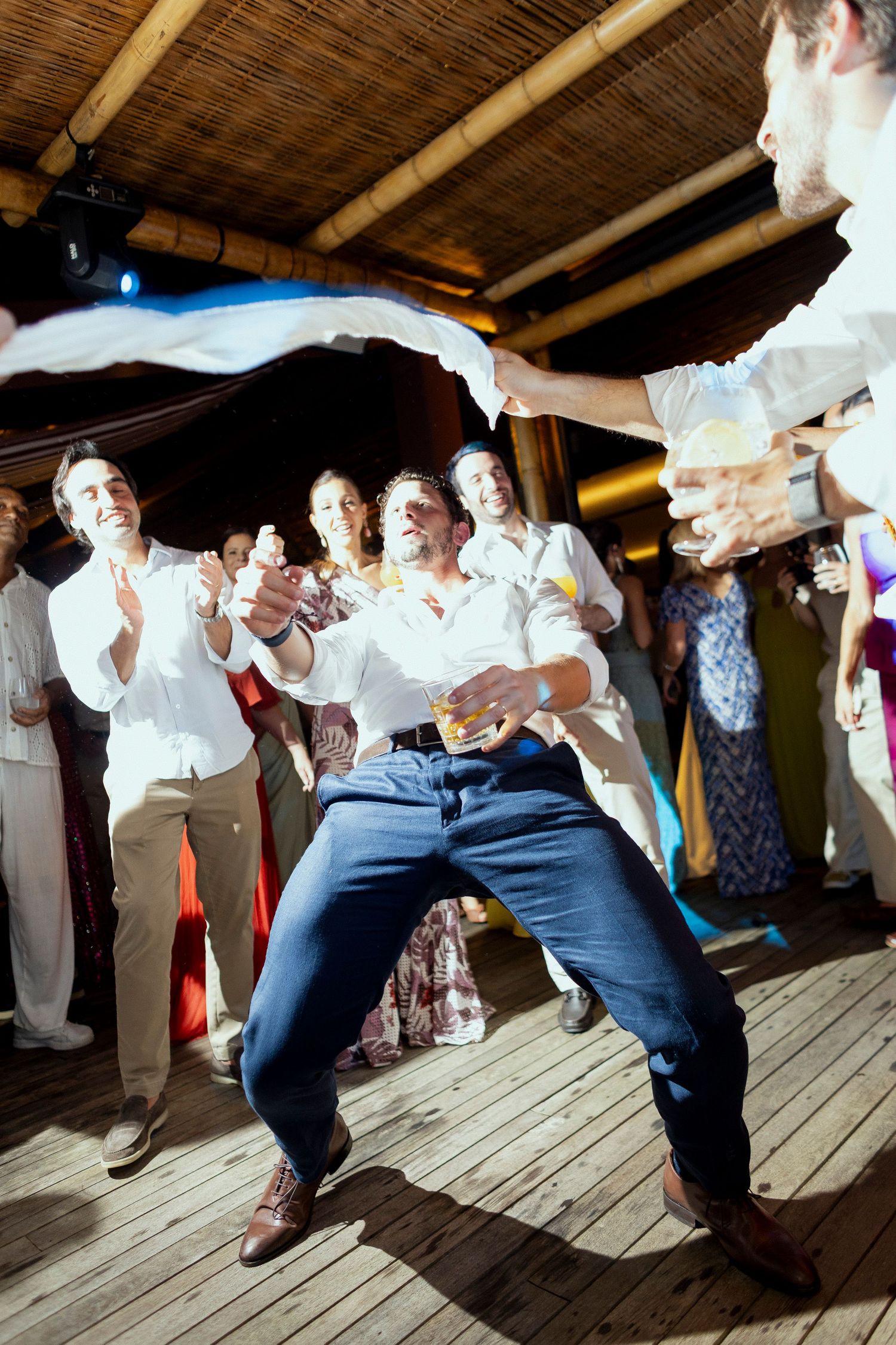 Man dancing energetically at a wedding reception party under bamboo ceiling while guests cheer around him.