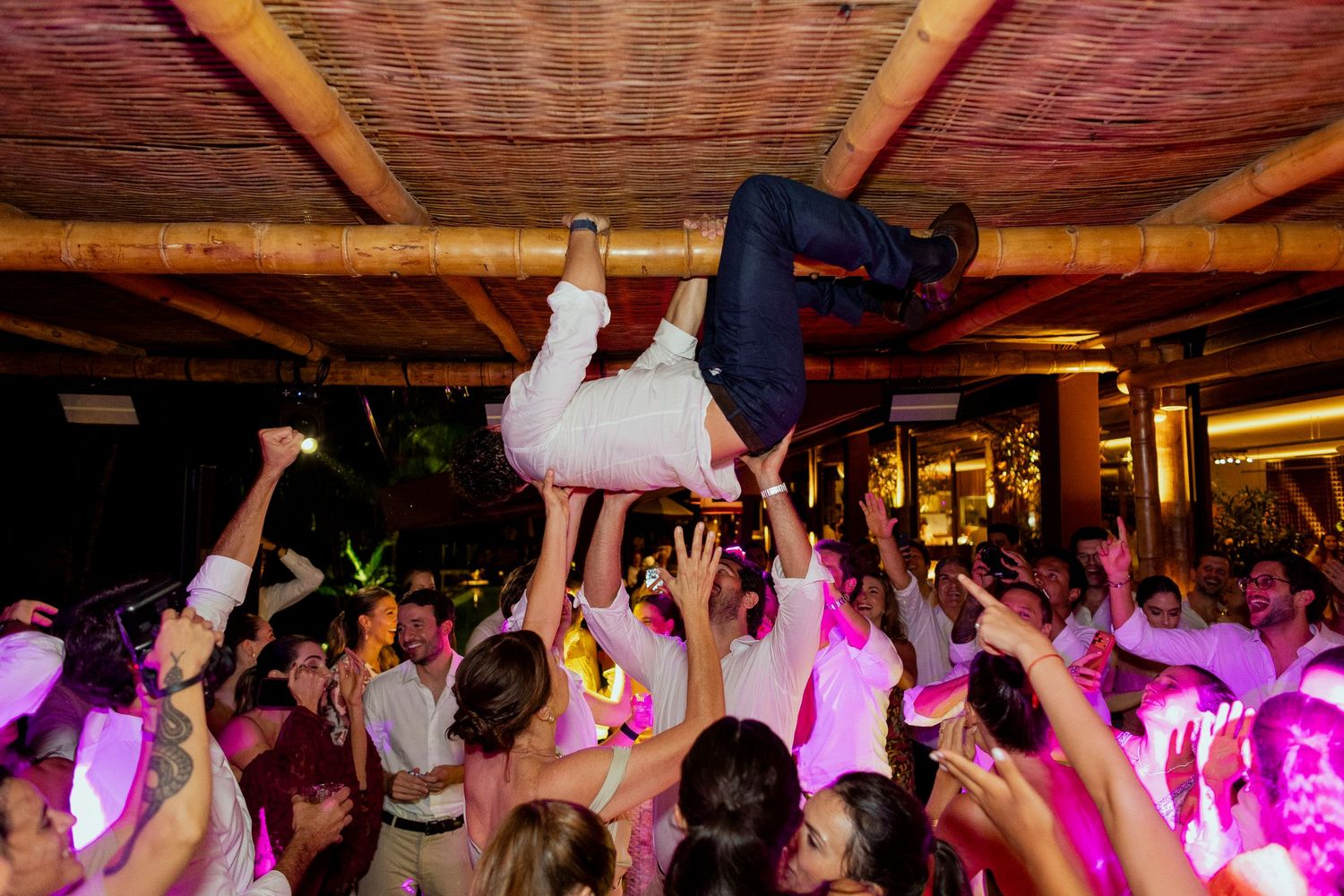 Wedding guests crowd surfing a person at a lively reception party with pink lighting.