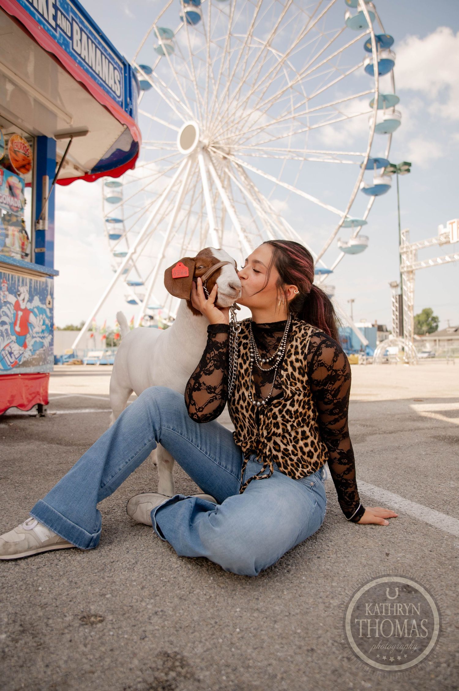 A young woman in leopard print top and jeans sits on pavement kissing a dog at a carnival with a Ferris wheel behind her.