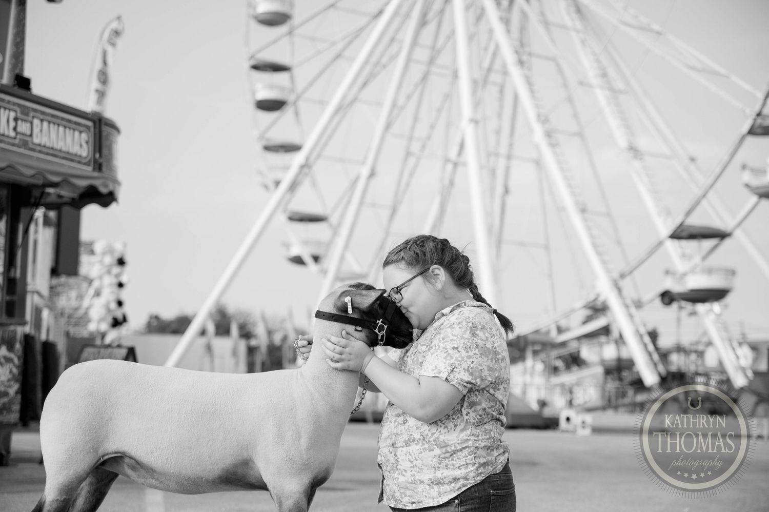 Girl bonding with a white sheep at a county fair with a Ferris wheel in the background, black and white photo.