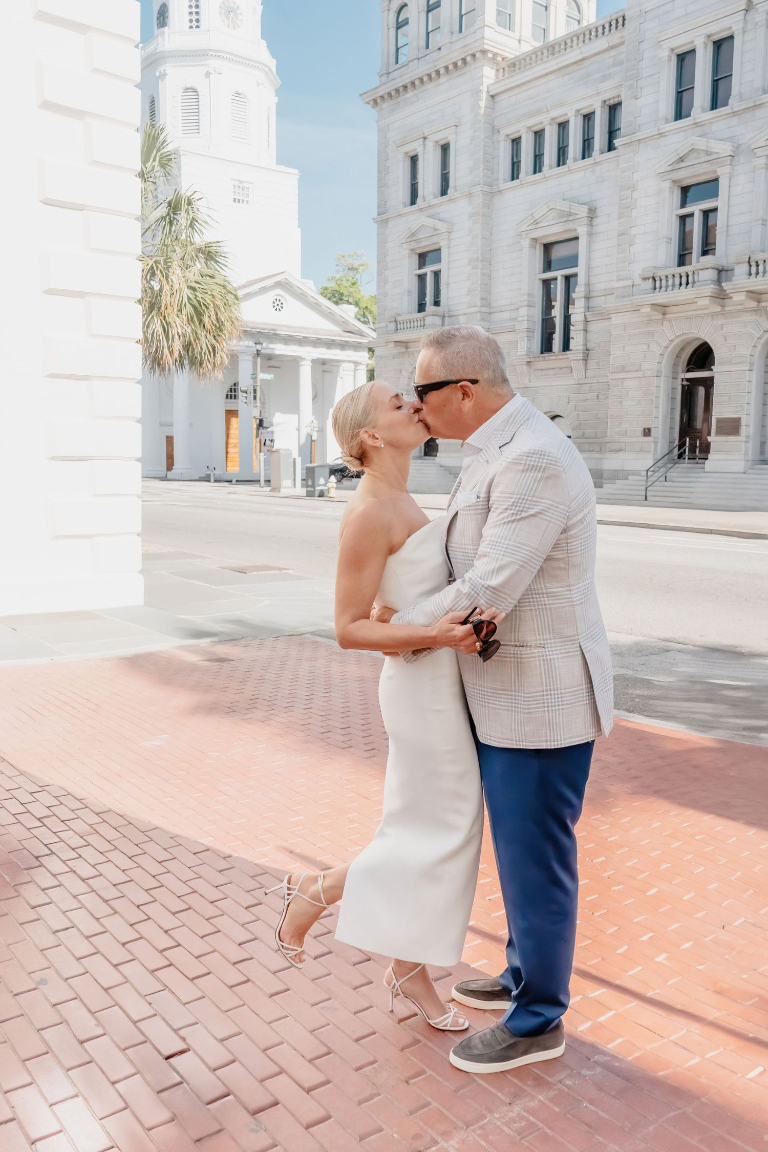 Couple sharing a kiss in white wedding attire on a sunny brick plaza with historic buildings backdrop.