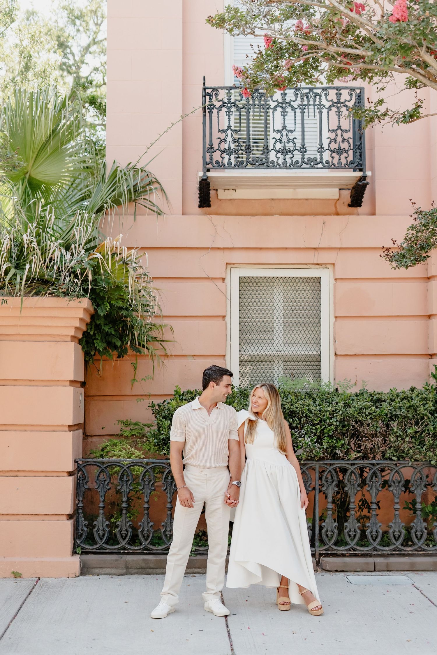 Couple holding hands in front of a pink building with wrought iron balcony and lush greenery.
