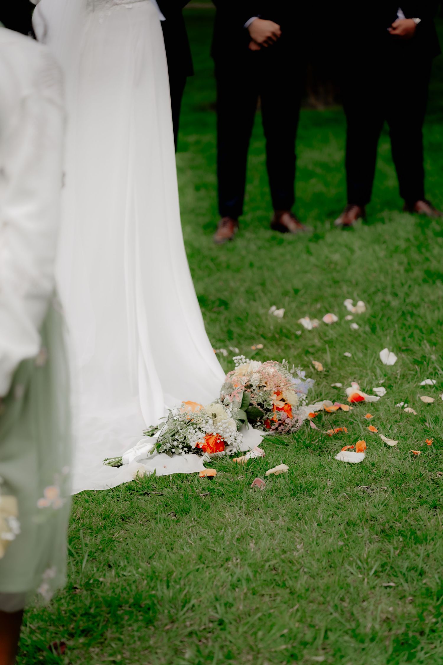 Bridal bouquet of orange and white flowers resting on green grass beside a flowing white wedding gown train.