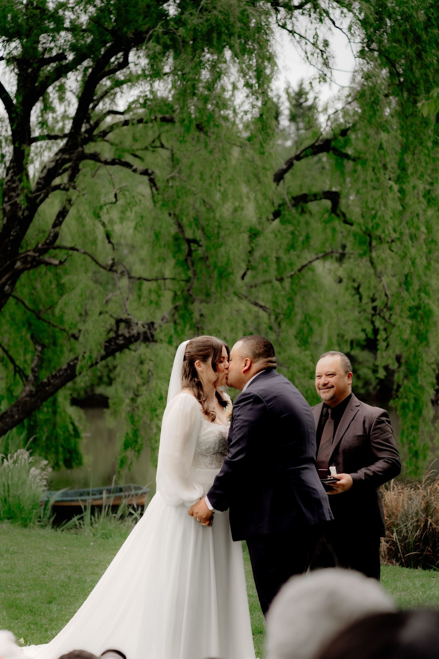 Bride and groom share first kiss at outdoor wedding ceremony under a weeping willow tree with officiant smiling beside them.