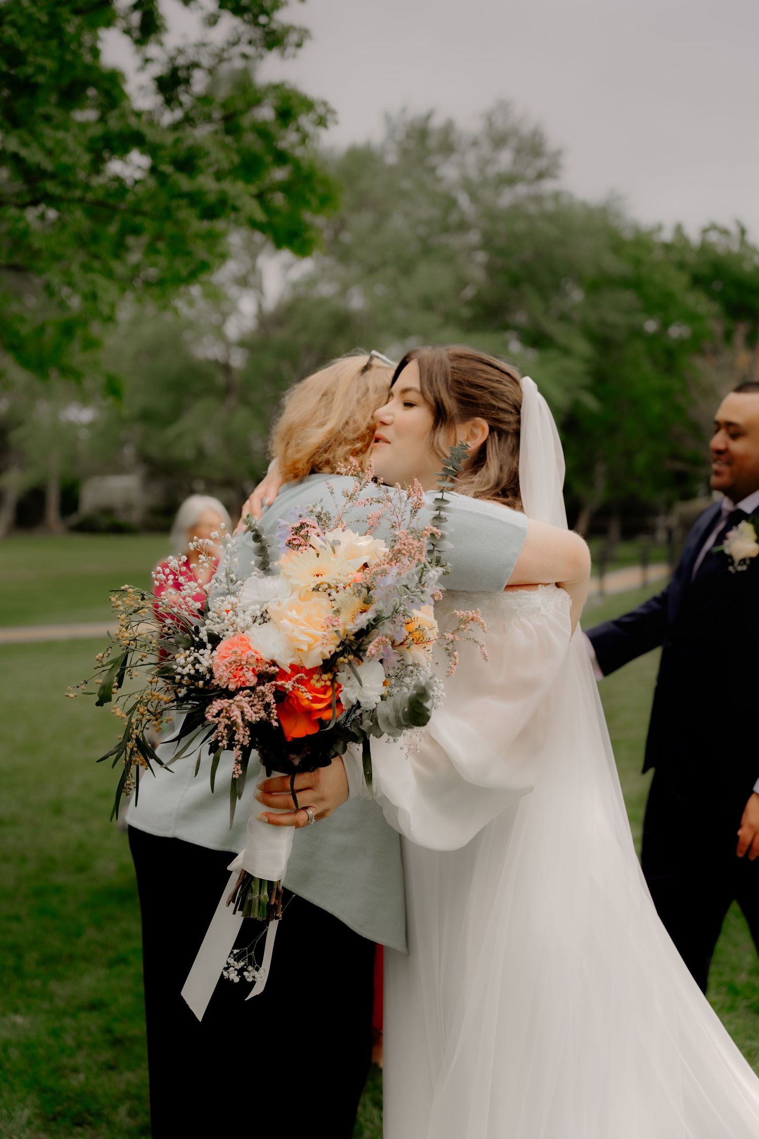 Bride in white gown and veil hugging a guest while holding a colorful bouquet at an outdoor wedding.