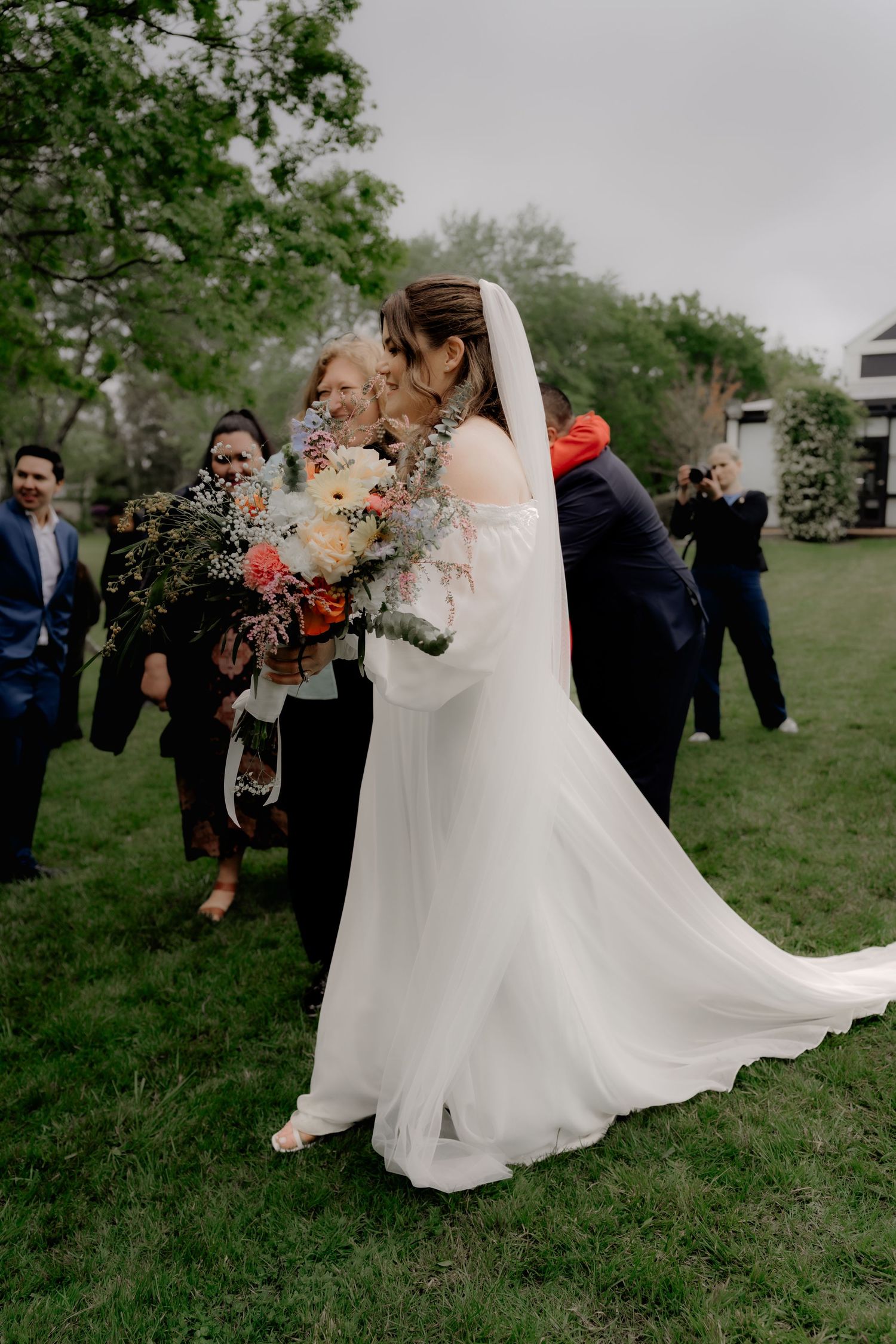 Bride in elegant white gown with veil holding colorful bouquet surrounded by wedding guests outdoors.