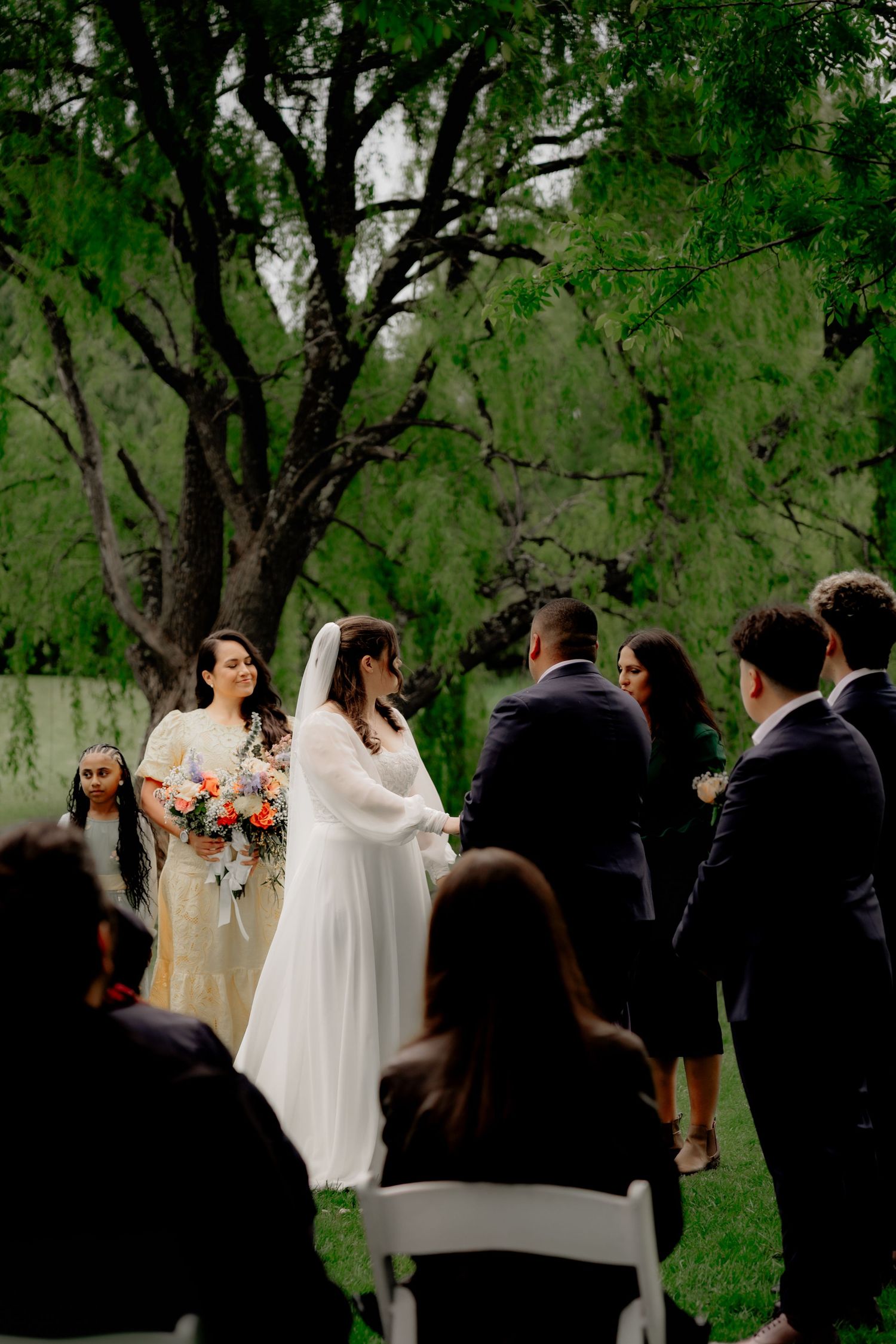 Bride in white gown and veil exchanges vows outdoors under lush green trees with guests seated nearby.