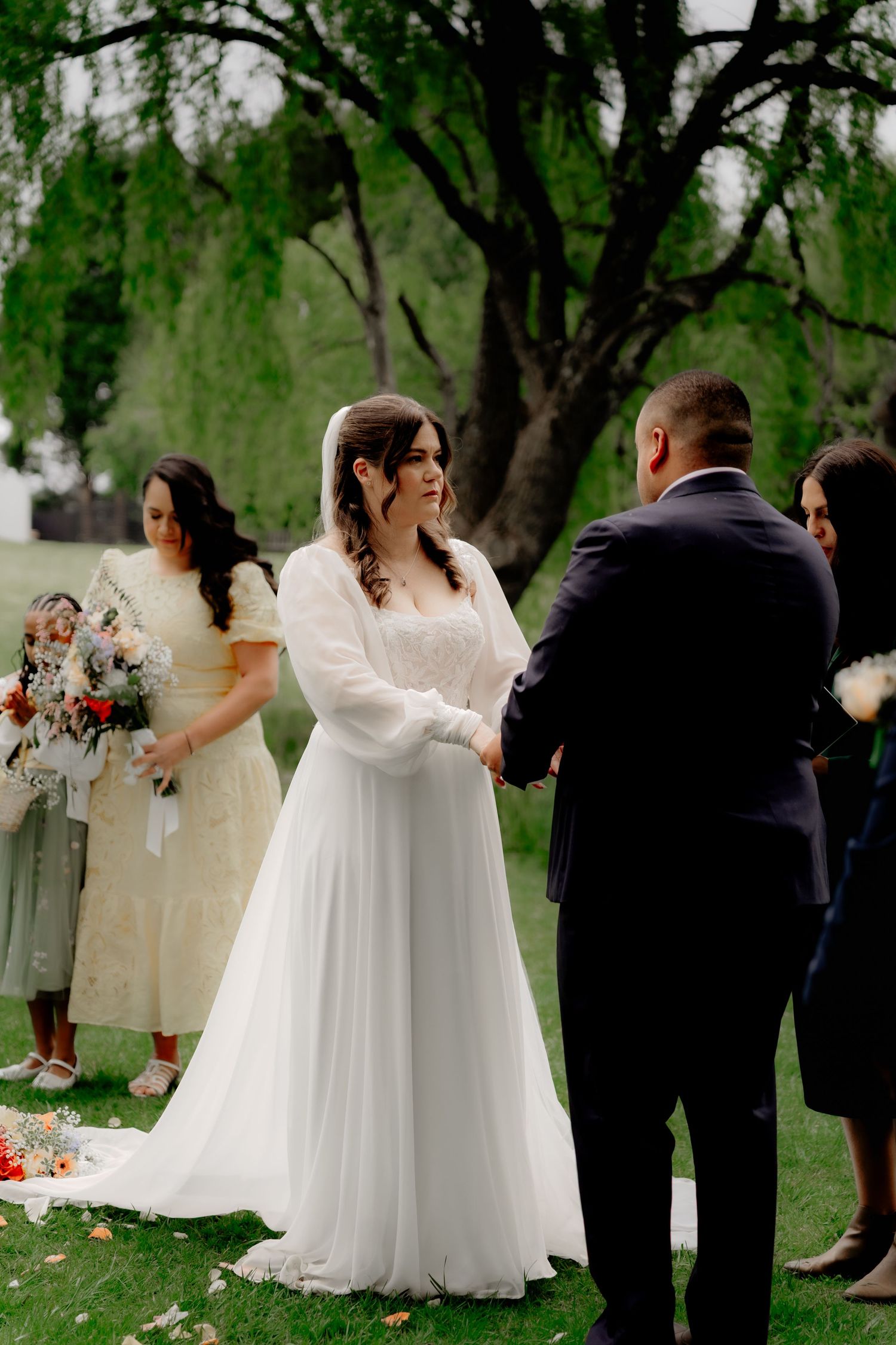 Bride in white gown and veil holds hands with groom during outdoor wedding ceremony under a green tree.