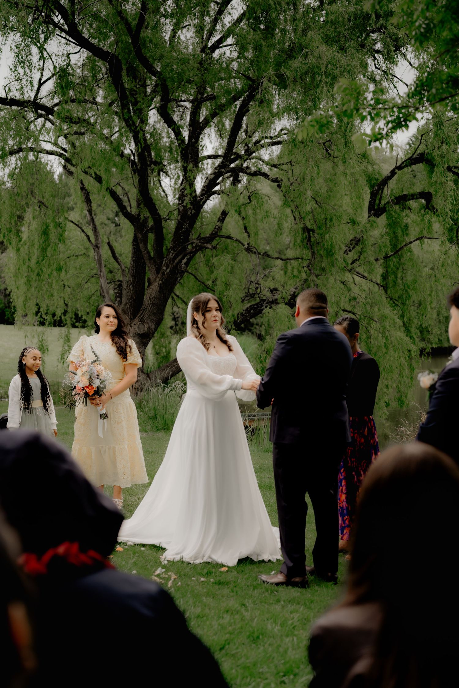 Bride in white gown exchanges vows outdoors under a willow tree, with bridesmaids holding bouquets nearby.