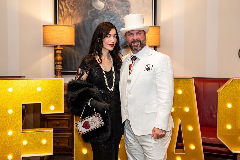 Elegant couple posing in formal attire beside illuminated marquee letters at a glamorous event.