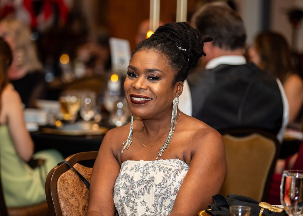 Elegant woman in strapless beaded gown smiling at formal gala event with guests in background.