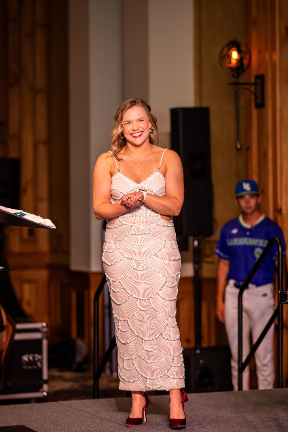 Smiling woman in elegant beaded white dress and red heels standing on stage at a formal event.