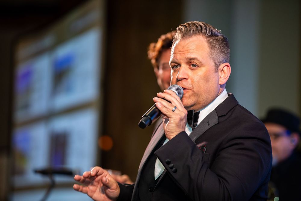 Professional male speaker holding microphone at corporate event with presentation screens in background.
