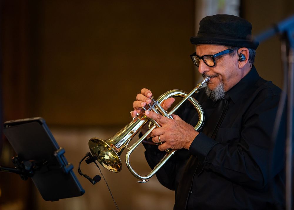 A jazz musician in a black hat and glasses plays a brass trumpet on stage under warm lighting.