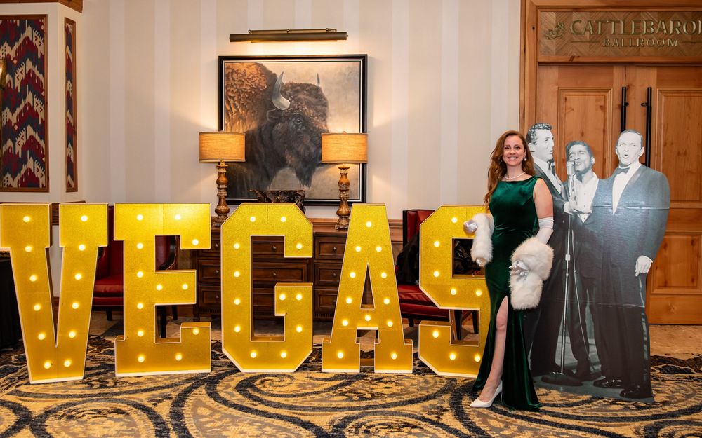 Woman in green gown poses beside lit VEGAS marquee letters and celebrity cardboard cutouts in elegant venue.