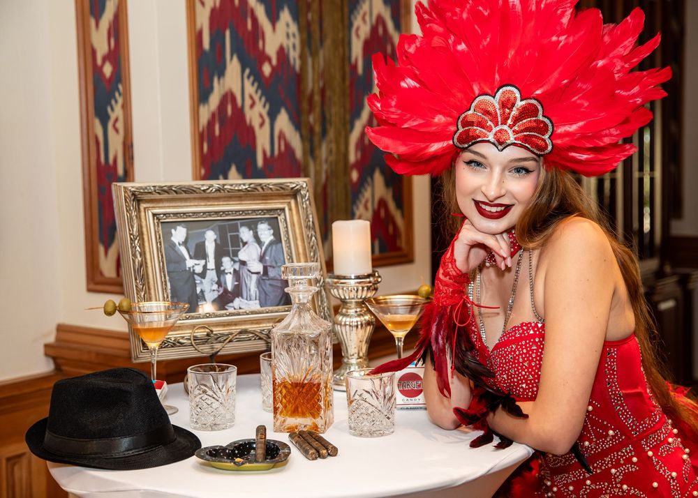 Woman in red showgirl costume with feather headdress poses at vintage-styled table with candles and framed photo.