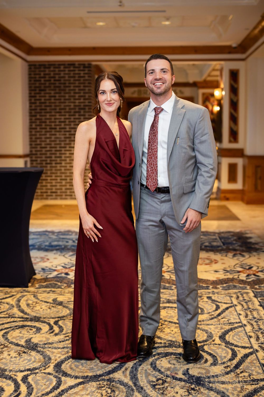 Couple posing at formal event, woman in burgundy gown and man in gray suit with red tie, elegant ballroom.