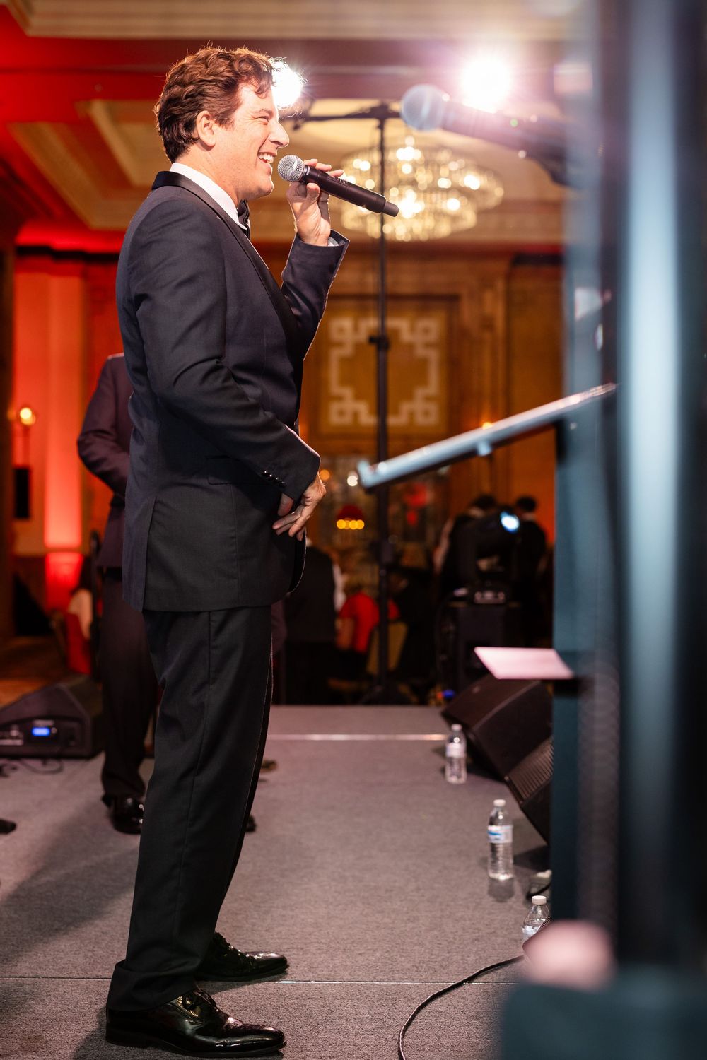 Man in dark suit singing into microphone on stage at elegant event with chandelier and red lighting.