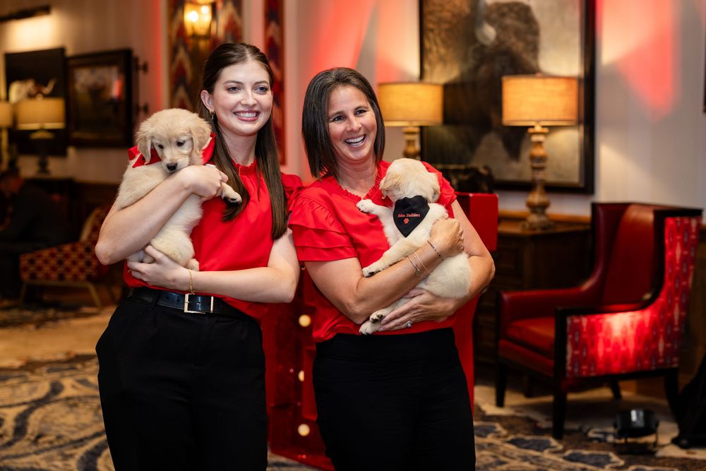 Two women in red tops holding adorable puppies smile in an elegant hotel lobby setting.