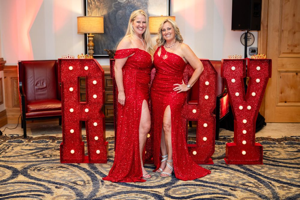 Two women in red sequin gowns pose in front of marquee letter lights at a formal event.