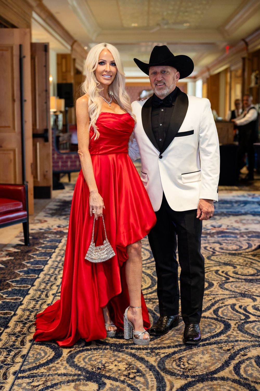 Elegant couple posing in hallway, woman in red satin gown, man in white tuxedo jacket and black cowboy hat.