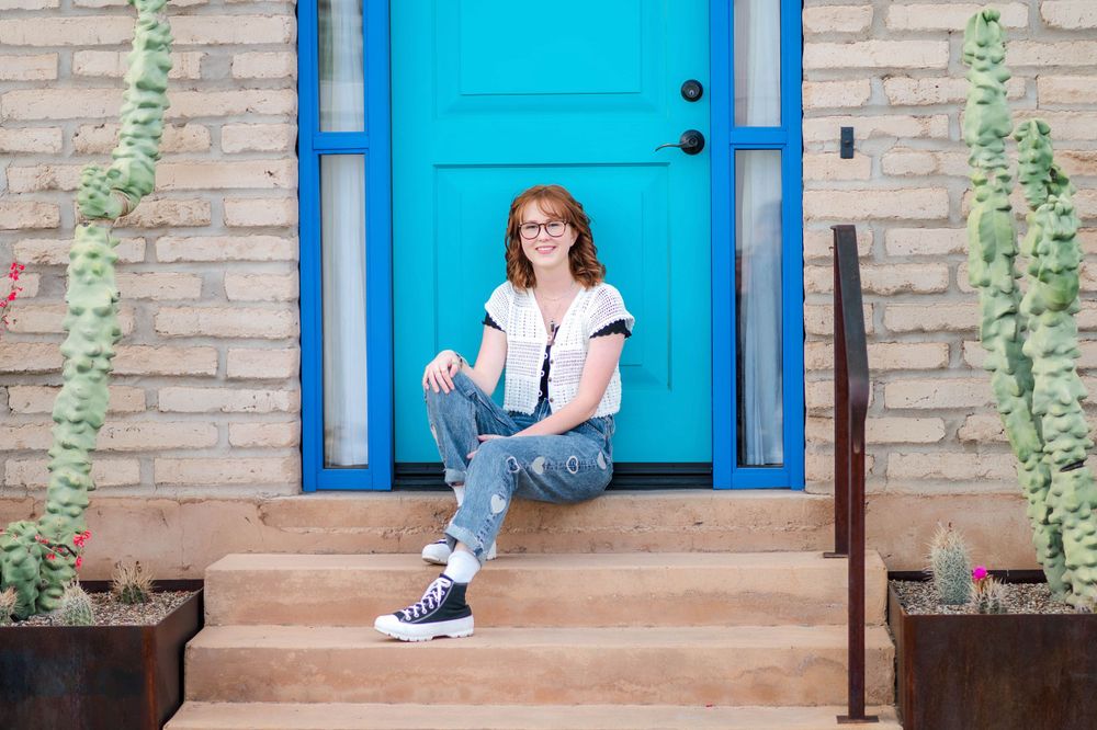 Redheaded teen in casual outfit sitting on steps in front of a vibrant blue door.