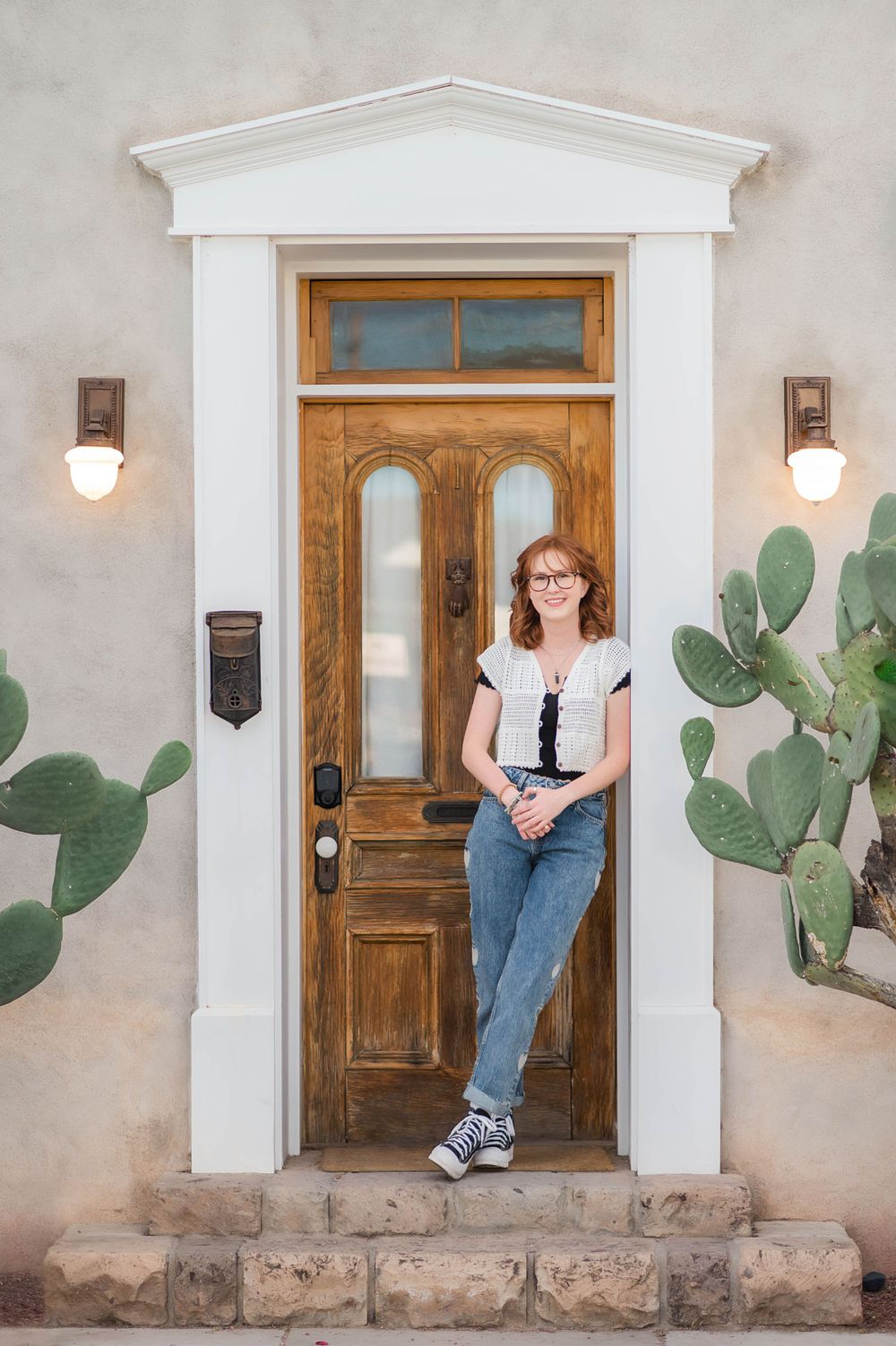 Teen girl in jeans leaning against a wooden door flanked by tall prickly pear cacti.