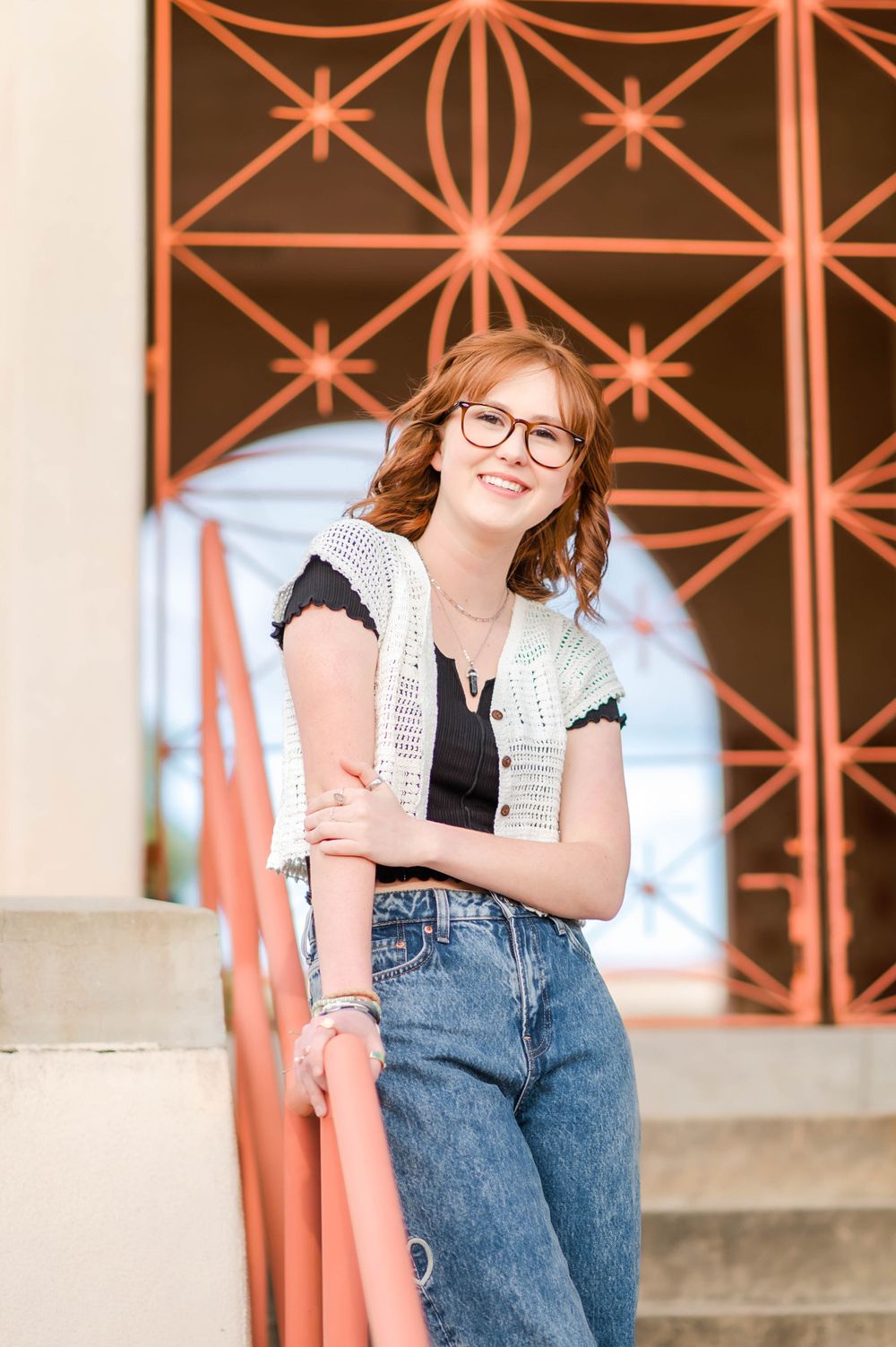 Smiling teen with glasses leaning on an orange railing in front of a decorative metal wall.