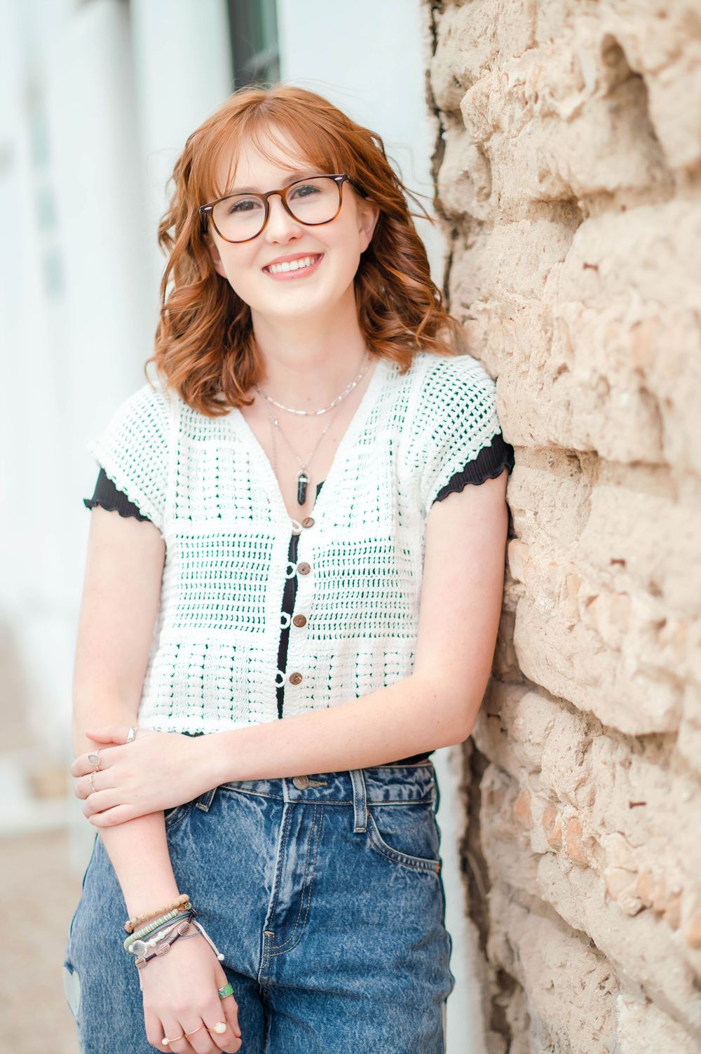 Smiling redhead teen in crochet top and jeans leaning against a stone wall outdoors.