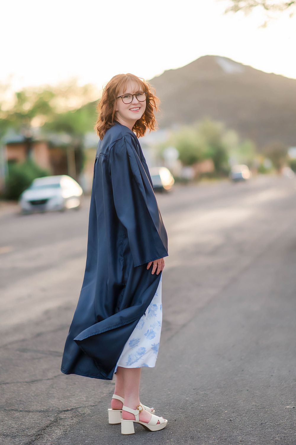 Graduate in navy cap and gown smiling on a street with mountain backdrop at sunset.