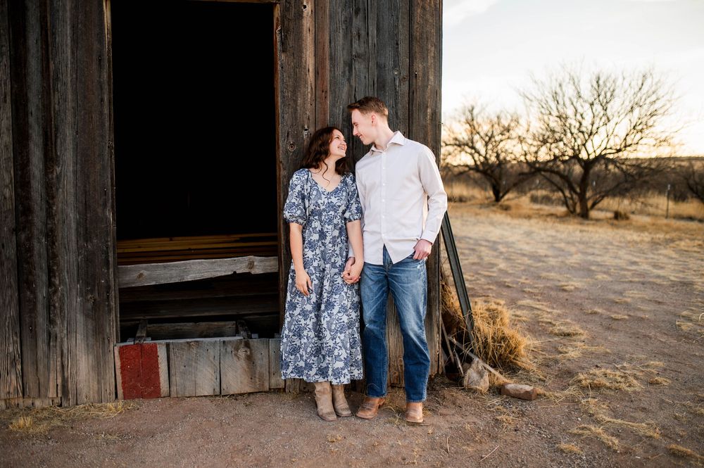 Couple stands together in a vintage barn doorway in a desert landscape at sunset.