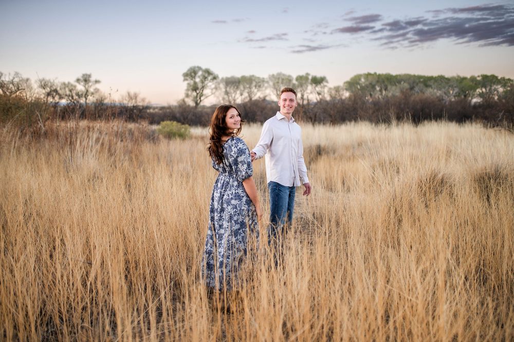 Couple holds hands walking through tall golden grass field at twilight in nature.