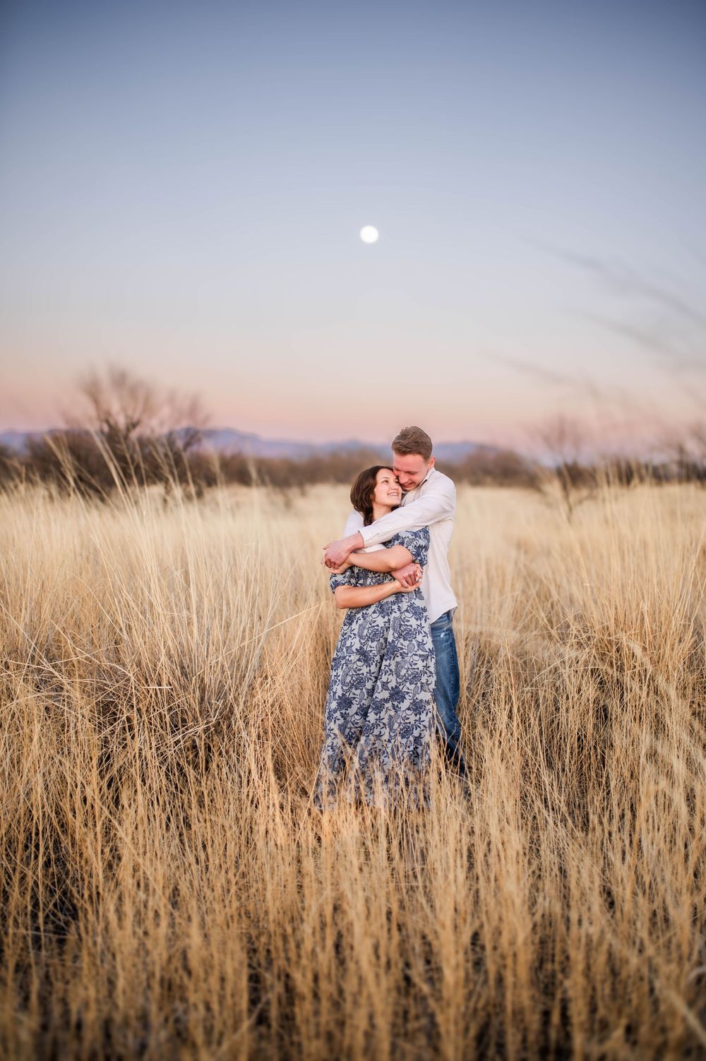 Couple embraces romantically in golden wheat field under a full moon at dusk.