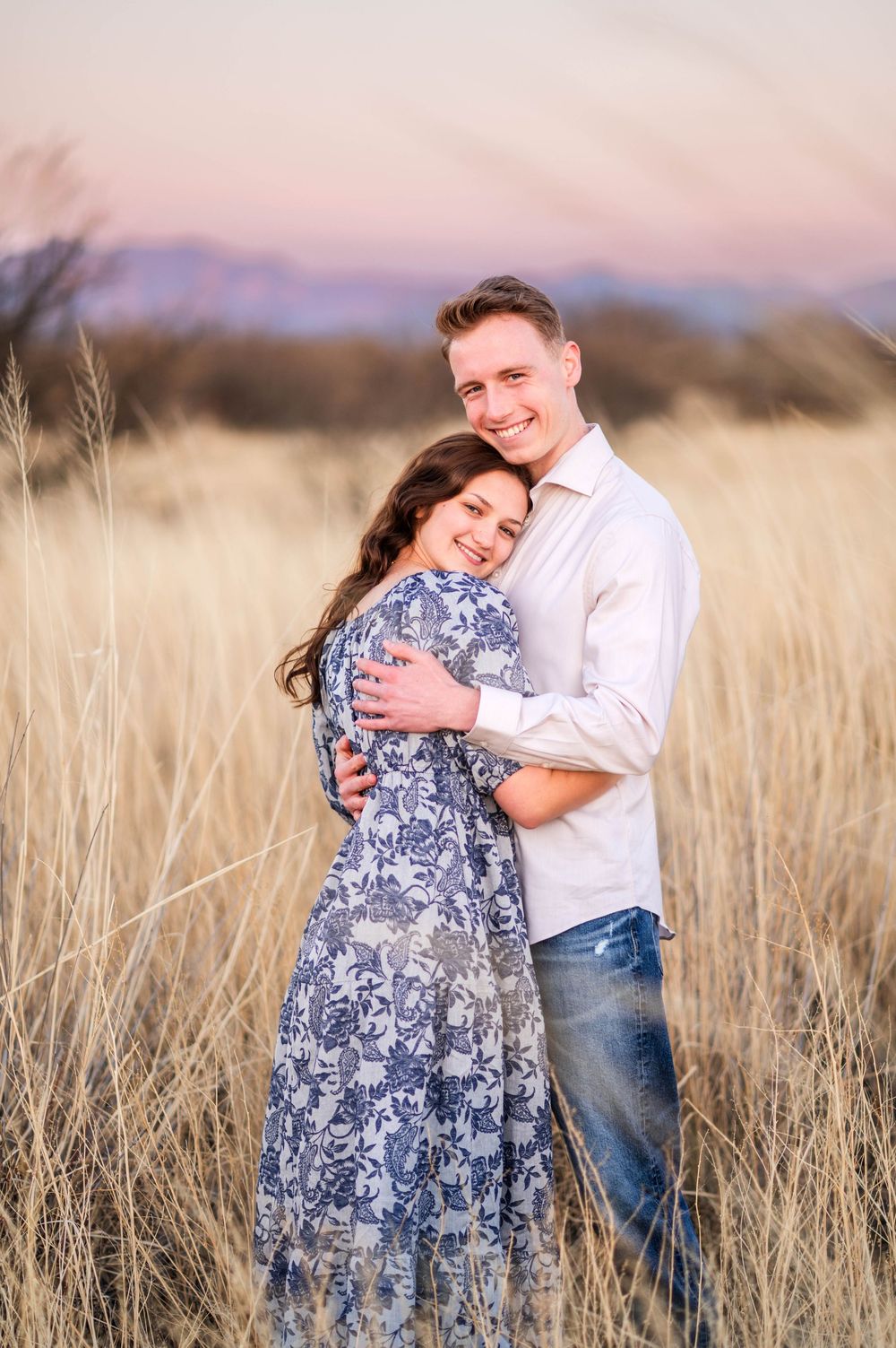 Smiling couple hugs warmly in tall golden grass field during a beautiful sunset.