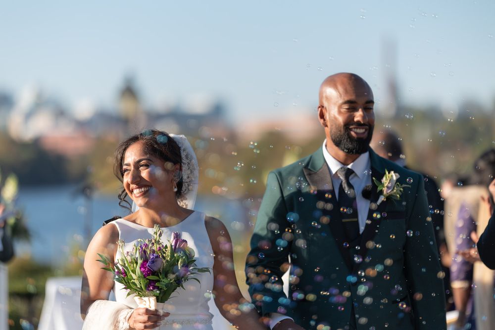 Newlywed couple walks through bubbles after their wedding ceremony, bride holding a colorful bouquet outdoors.