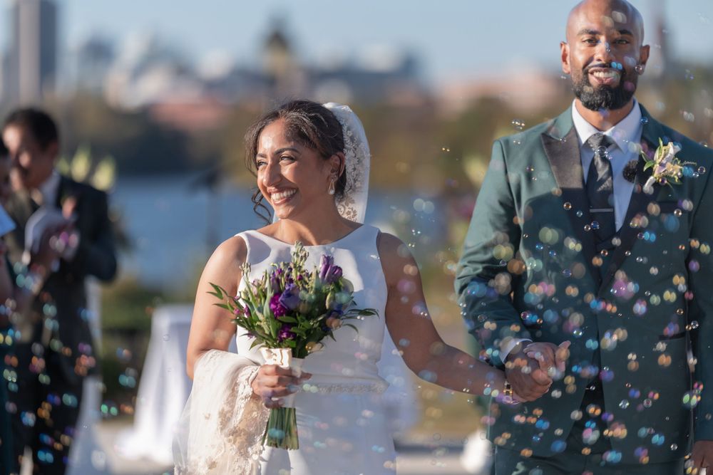 Joyful interracial couple walks through bubbles after outdoor wedding ceremony, bride holding colorful bouquet in white gown.