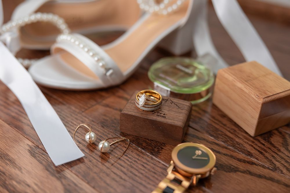 Bridal accessories flat lay with pearl heels, gold rings, earrings, perfume, and a watch on wood floor.