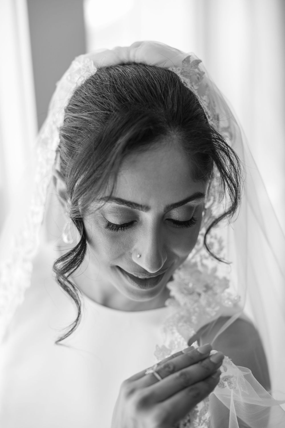 Black and white portrait of a smiling bride in a lace veil looking down, holding her veil delicately.