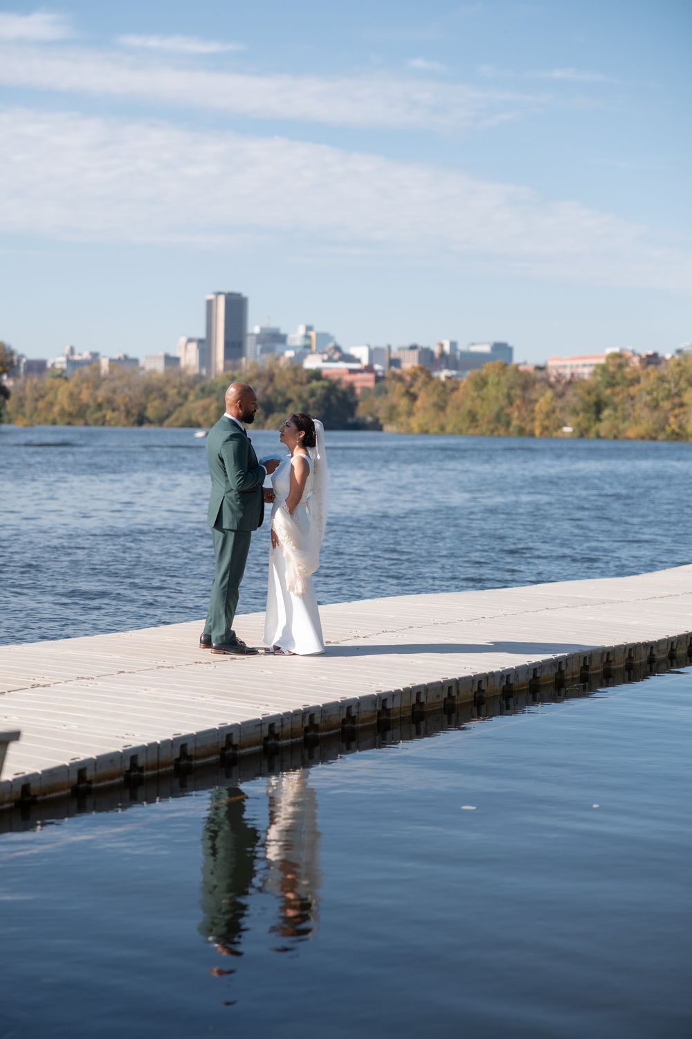 Bride and groom standing on a dock overlooking a river with a city skyline backdrop on a sunny day.