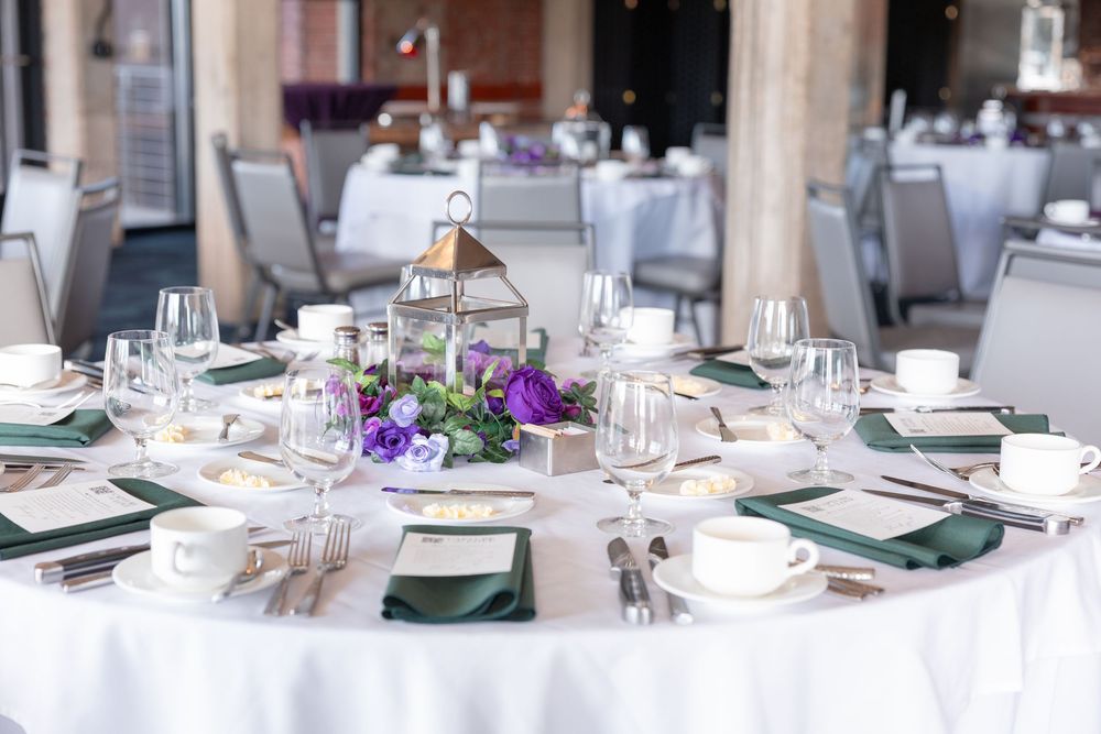 Elegant wedding reception table with white linen, green napkins, purple floral centerpiece, and gold lantern accent.