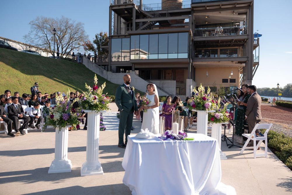 Outdoor wedding ceremony with couple at altar, floral arrangements, and guests seated near a modern building.