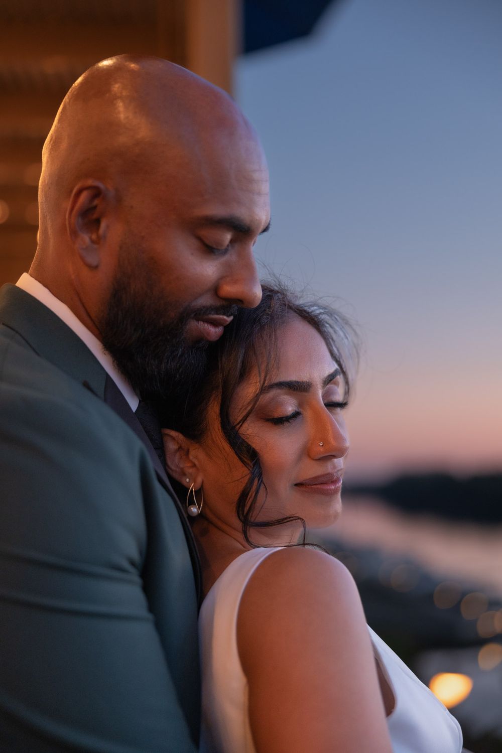 A couple shares a tender embrace at sunset, the groom in a gray suit holding his bride in a white dress.